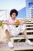 Black woman texting with her smartphone, sitting on steps of a modern building. photo