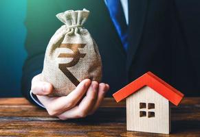 Man holds out a indian rupee bag near the house. Property appraisal. Favorable terms and conditions, low interest rate. Bank approval for issuing a mortgage loan. Home purchase, invest in real estate. photo