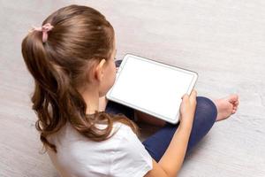 Little girl sitting on the floor and holding a white tablet with isolated screen for promoting video games, websites or apps photo