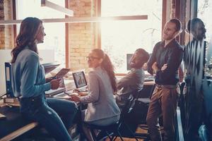 Group of young modern people in smart casual wear communicating and using modern technologies while working in the office photo