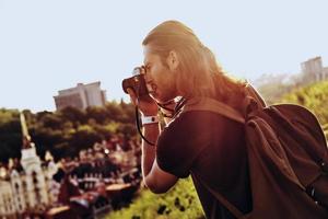 Nice shot. Young man in casual clothing photographing the view while standing on the hill outdoors photo