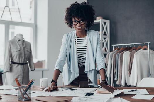 listo para hacer tu sueño realidad. hermosa joven africana sonriendo y mirando la cámara mientras se apoya en el escritorio en el taller con ropa colgada en el fondo foto