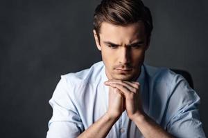 Calm and handsome. Portrait of handsome young man keeping hands clasped and looking thoughtful while sitting against black background photo