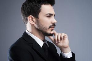 Thinking about solutions. Side view of thoughtful young man in formalwear looking away and holding hand on chin while standing against grey background photo