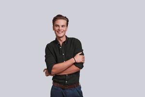 Young and confident. Good looking young man keeping arms crossed and looking away while standing against grey background photo