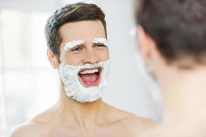 Shaving with fun. Rear view of playful young man with shaving cream on his face smiling while standing in front of the mirror photo