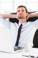 Day dreaming in office. Handsome young man in shirt and tie holding hands behind head and smiling while sitting at his working place photo