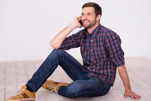 Good talk with friend. Handsome young man talking on the mobile phone and smiling at camera while sitting on hardwood floor photo