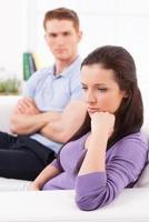 Problems with relationship. Depressed young woman holding hand on chin and looking away while man sitting behind her on the couch and keeping arms crossed photo