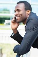 buena charla de negocios. vista lateral de un joven africano feliz con ropa formal hablando por teléfono móvil y sonriendo mientras está de pie al aire libre foto