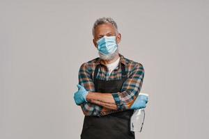 Senior man in apron and protective face mask looking at camera while standing against gray background photo
