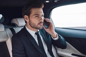 tomando todo en serio. un joven apuesto con traje completo hablando por teléfono inteligente mientras está sentado en el auto foto