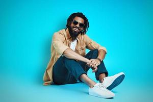 Trendy young African man in casual wear smiling and looking at camera while sitting against blue background photo