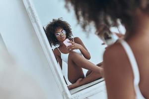 Her body is perfection. Reflection of beautiful young African woman in white lingerie taking selfie in the mirror while sitting on the floor at home photo