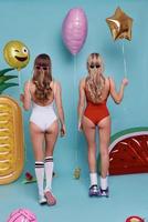 Ready for fun.  Full length rear view of two beautiful young women in swimwear wearing eyeglasses and holding balloons while standing against blue background photo