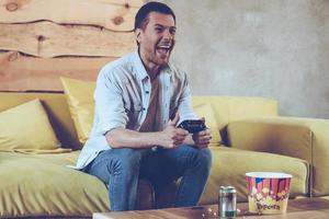 juegos siempre divertidos. alegre joven apuesto jugando videojuegos y luciendo emocionado mientras se sienta en el sofá en casa foto