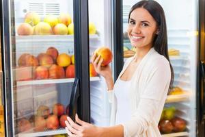 eligiendo la manzana más fresca. hermosa joven sonriente eligiendo manzanas del refrigerador en la tienda de comestibles foto