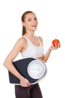 Leaving a healthy life. Side view of beautiful young woman in sports clothing holding weight scales and apple while standing isolated on white photo