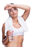 Refreshing after work out. Tired young woman holding bottle with water and touching her forehead with hand while standing isolated on white photo