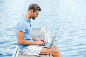 le gusta trabajar al aire libre. vista lateral de un apuesto joven con camisa de polo que trabaja en una laptop mientras se sienta en el muelle foto