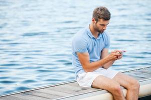 Checking messages from friends. Handsome young man in polo holding mobile phone and looking at it while sitting on quayside photo