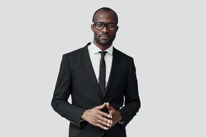 Confident young African man in formalwear looking at camera and keeping hands clasped while standing against grey background photo