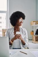 Beautiful young African woman using smart phone and smiling while sitting at her working place in office photo