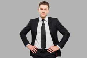 Young and successful. Confident young man in formalwear looking at camera while holding hands on hips and standing against grey background photo