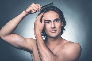 Everything should be perfect. Portrait of young shirtless man combing his hair with hairbrush and looking at camera while standing against grey background photo