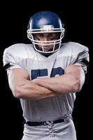 Confident in football player.  Portrait of American football player looking at camera and keeping arms crossed while standing against black background photo