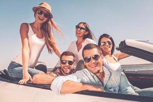Spending great time together. Group of young happy people enjoying road trip in their white convertible and smiling at camera photo