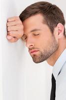 Sick and tired. Side view of depressed young man in shirt and tie leaning at the wall and keeping eyes closed photo