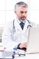 médico general médico de cabello gris maduro que trabaja en una computadora portátil mientras está sentado en su lugar de trabajo foto