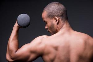 Making his body strong. Rear view of young shirtless African man exercising with dumbbell while standing against grey background photo