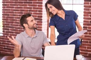 Creative collaboration. Cheerful young man sitting at his working place and gesturing while beautiful woman standing close to him and smiling photo