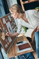 Taking notes. Thoughtful young woman writing something down on the adhesive note while standing in the workshop photo