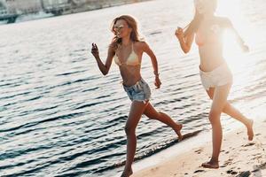 Young and full of energy. Two attractive young women in swimwear smiling while running on the beach photo