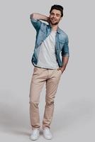 Trendy look. Full length of handsome young man in blue jeans shirt holding hand behind head and looking at camera while standing against grey background photo