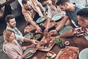 tiempo de pizza. vista superior de jóvenes con ropa informal recogiendo pizza y sonriendo mientras cenan foto