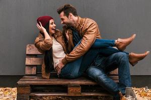 pareja juguetona. una pareja joven y juguetona que se divierten juntos mientras se sientan en el palet de madera junto con una pared gris en el fondo y hojas caídas en el suelo foto