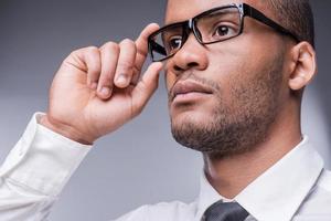 Confident businessman. Low angle view of thoughtful young African man in shirt and tie adjusting his eyeglasses and looking away while standing against grey background photo