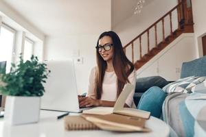 mujer joven atractiva con ropa informal usando una laptop y sonriendo mientras descansa en casa foto