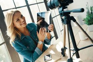 Beautiful young woman in casual clothing pointing tablet and smiling while sitting in front of digital camera photo