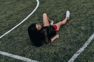 simplemente siendo ella misma. bella joven vestida con ropa deportiva manteniendo los ojos cerrados mientras se acuesta en el campo de fútbol foto