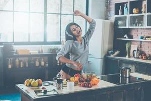 disfrutando de un nuevo día. hermosa joven mujer de raza mixta en auriculares cocinando ensalada y bailando mientras está de pie en la cocina en casa foto