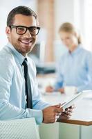 Confident business experts. Cheerful young businessman in formalwear holding digital tablet and looking at camera while his female colleague working in the background photo