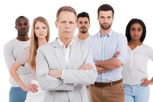Leading his team to success. Confident mature man in smart casual wear keeping arms crossed and looking at camera while group of young people standing behind him and against white background photo