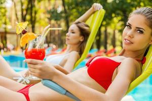 nos encanta pasar tiempo junto a la piscina. vista lateral de dos mujeres jóvenes atractivas en bikini bebiendo cócteles mientras se relaja en una tumbona cerca de la piscina foto