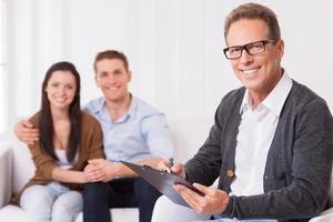 Confident family psychologist. Confident psychiatrist writing something in clipboard and smiling while cheerful couple sitting in the background and holding hands photo