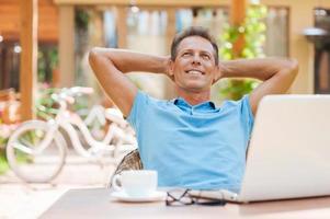 Happy day dreamer. Relaxed mature man holding hands behind head and smiling while sitting at the table outdoors with laptop on it photo